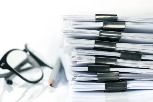 A close-up of stacked office work documents with a pen and glasses.