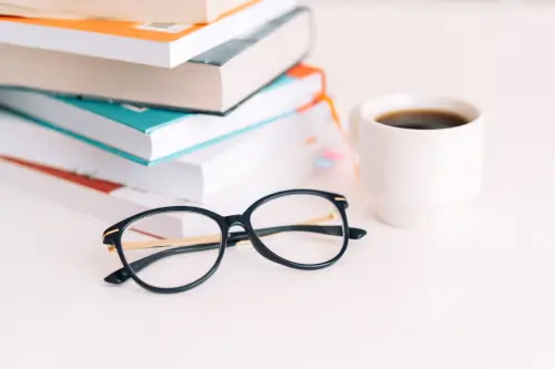 A stack of books, along with glasses and a cup of coffee, rests on a white table.